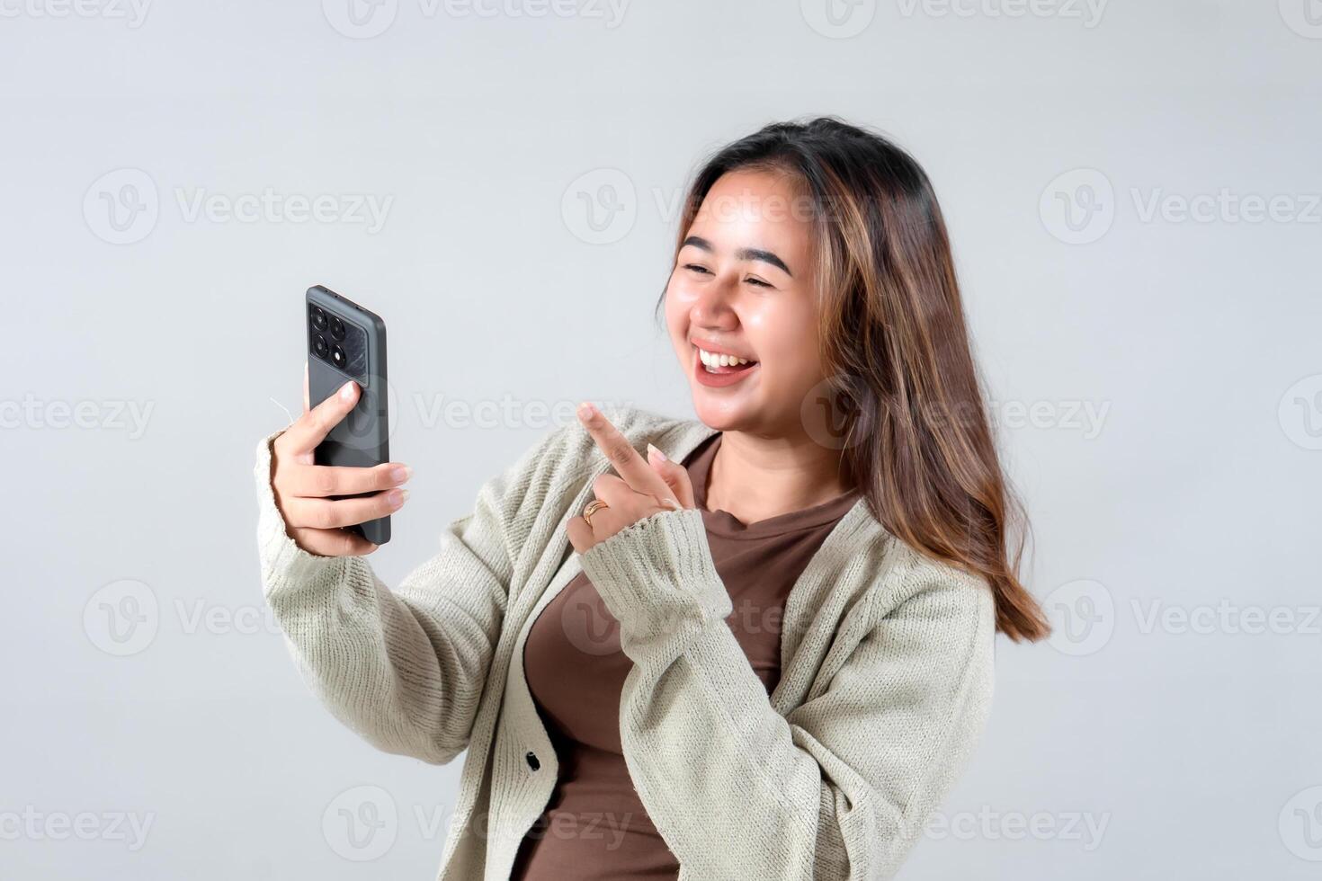 A young woman smiles and points at her smartphone, expressing joy and excitement while engaging with her device in a neutral background. photo