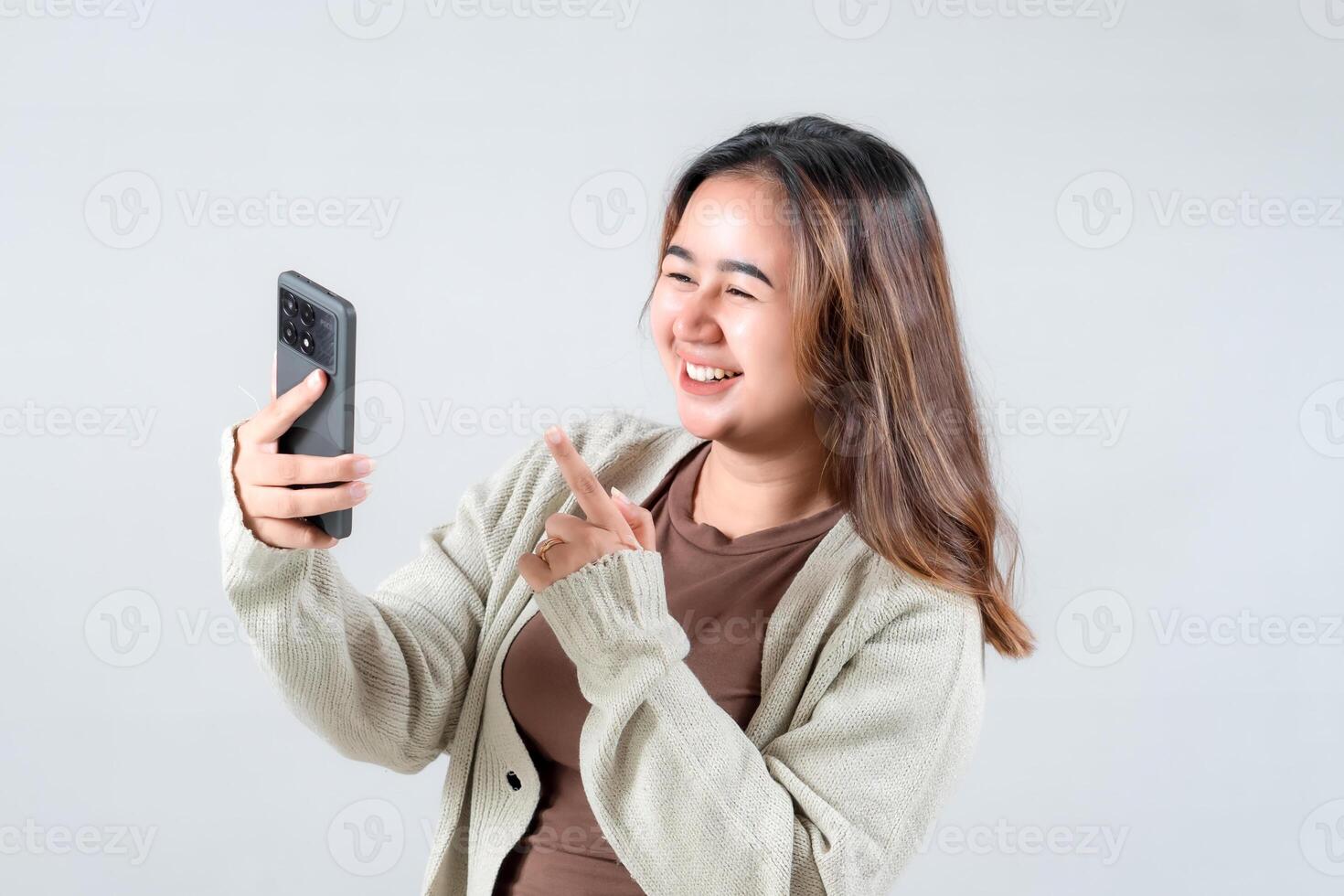 Young woman smiles and points at her smartphone, expressing joy and excitement while engaging with her device in a neutral background. photo
