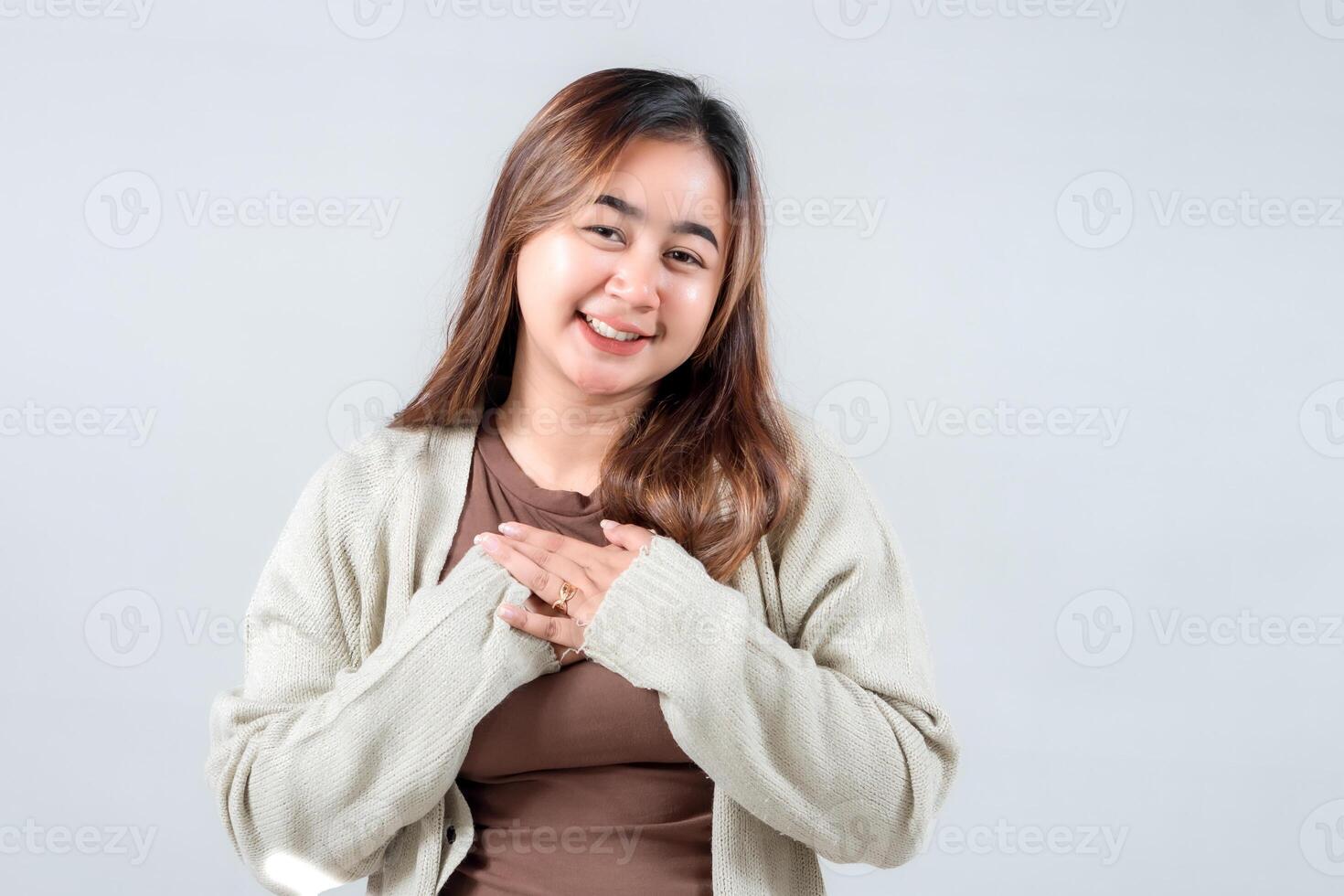 young woman with a warm expression, wearing a cozy sweater, holds her hands close to her heart, radiating kindness and positivity against a neutral backdrop. photo