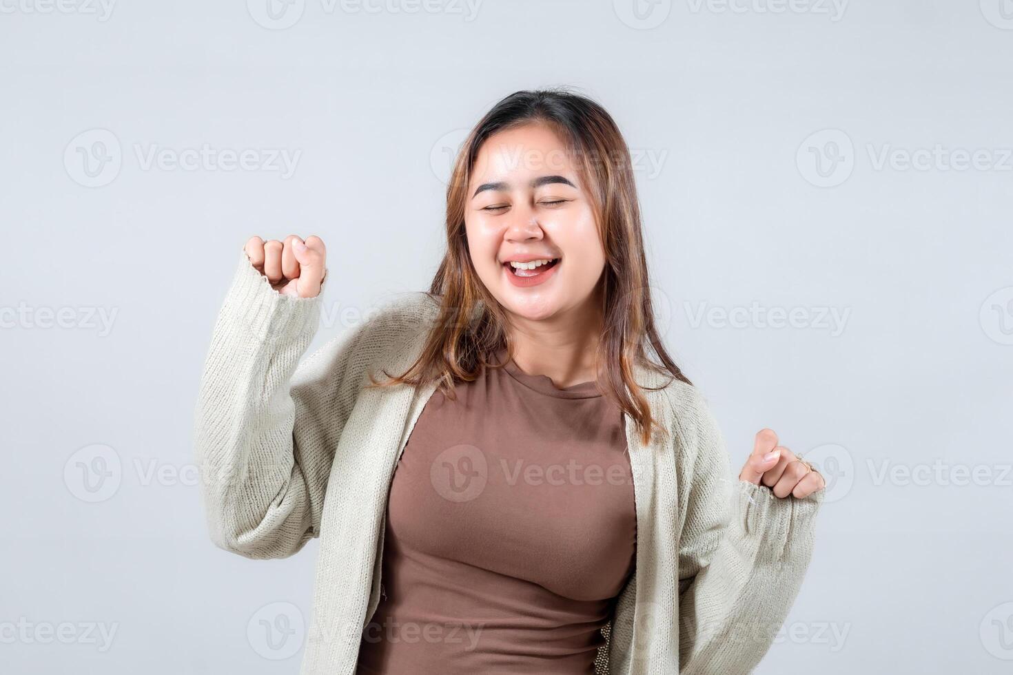 A young woman in a cozy sweater dances with joy, her eyes closed and a bright smile on her face, expressing happiness and carefree spirit against a neutral background. photo