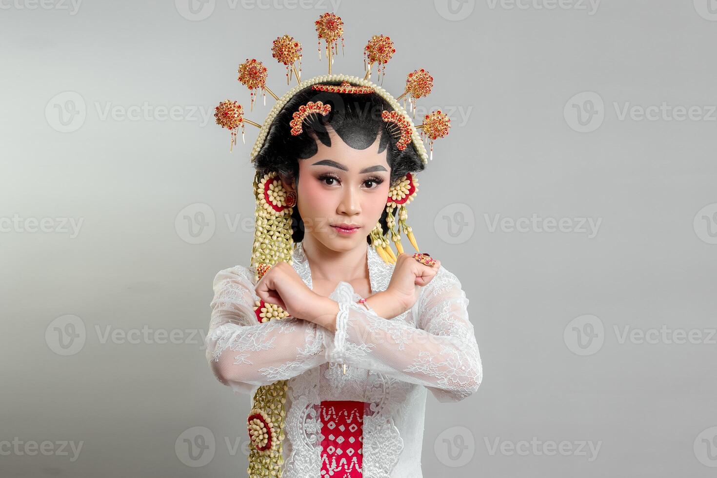 A Indonesian bride in Solo Putri attire, wearing an ornate headdress and lace kebaya. She crosses her arms in an X gesture with a determined expression, exuding strength and elegance. photo