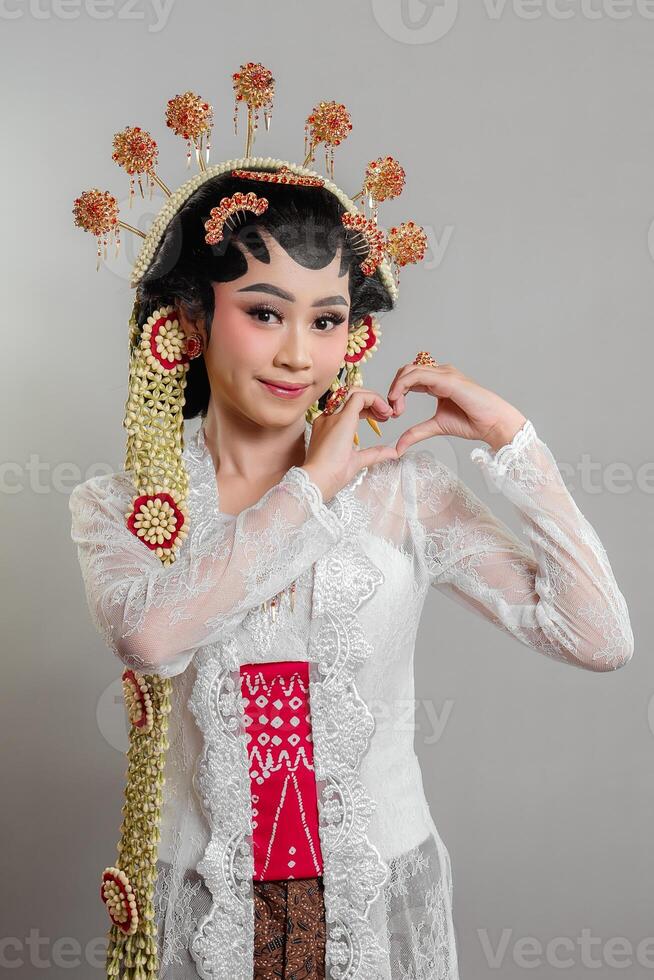 A Indonesian bride in Solo Putri attire, wearing a lace kebaya, intricate headdress, and jasmine garlands. She smiles sweetly while forming a heart shape with her hands. photo