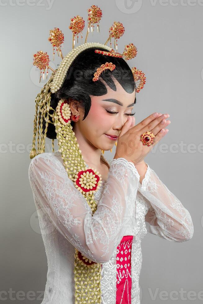 An Indonesian bride in Solo Putri attire stands gracefully with a soft smile hands in a respectful greeting. She wears a golden headdress, jasmine garland, and a white lace kebaya. photo