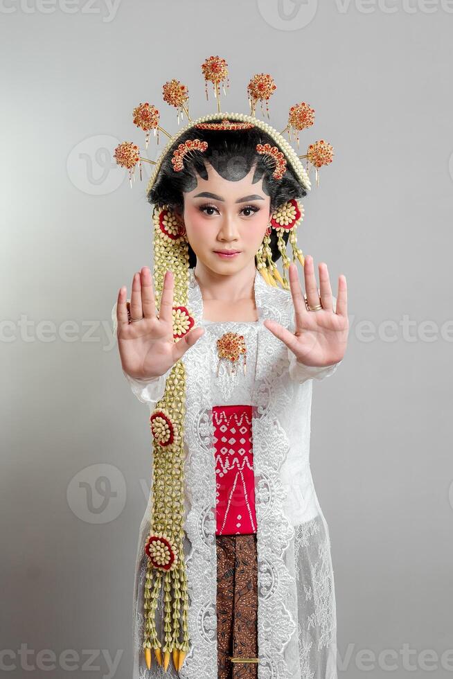Indonesian bride in Solo Putri attire, wearing an ornate headdress and lace kebaya. She raises both hands in a stop gesture with a serious expression, exuding elegance and grace. photo