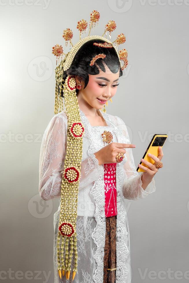 Indonesian bride in Solo Putri attire smiles while using a smartphone. She wears a golden headdress, intricate jewelry, a white lace kebaya, and traditional Javanese accessories. photo