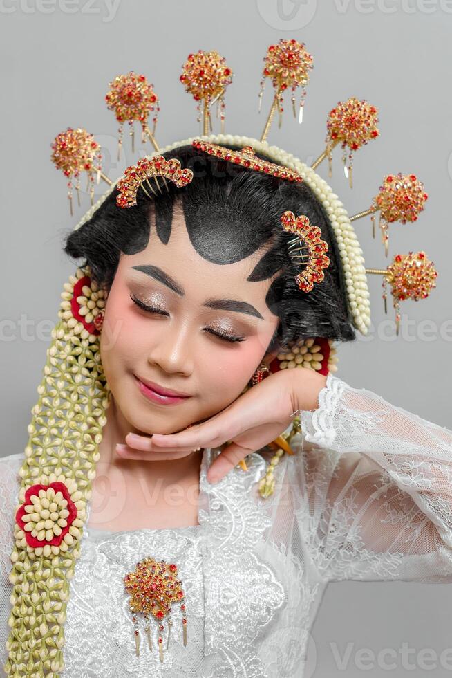 Indonesian bride in Solo Putri attire poses gracefully, resting her chin on her hand. She wears an intricate golden headdress, jasmine garland, and elegant lace kebaya. photo