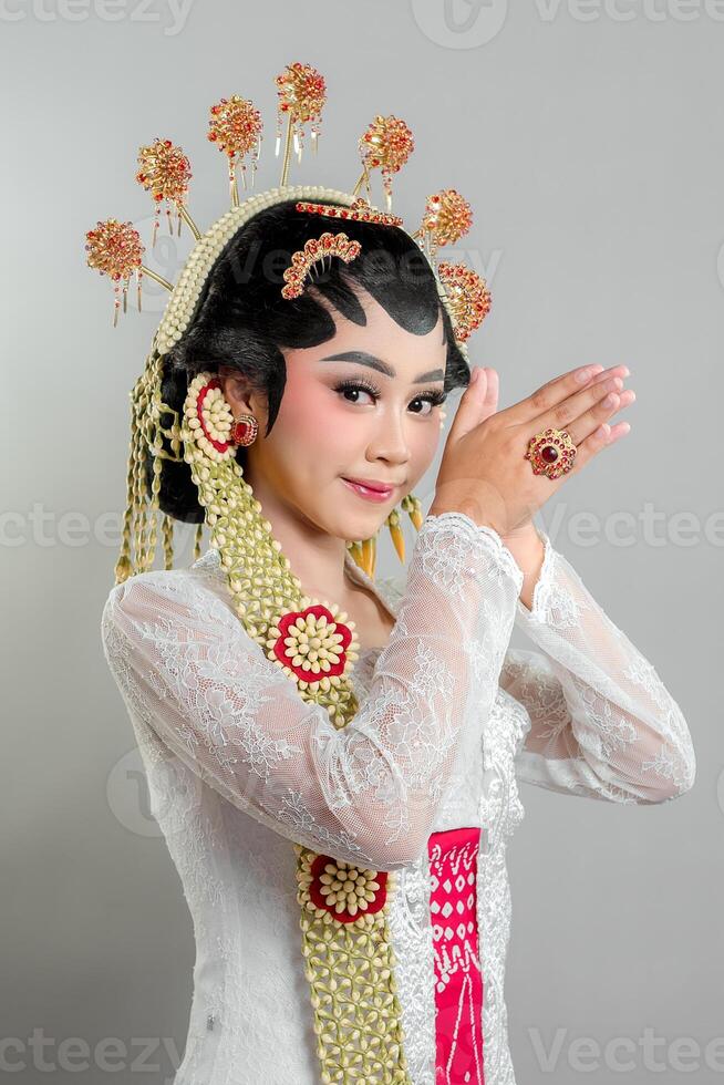 Indonesian bride in Solo Putri attire stands gracefully with a soft smile, hands in a respectful greeting. She wears a golden headdress, jasmine garland, and a white lace kebaya. photo