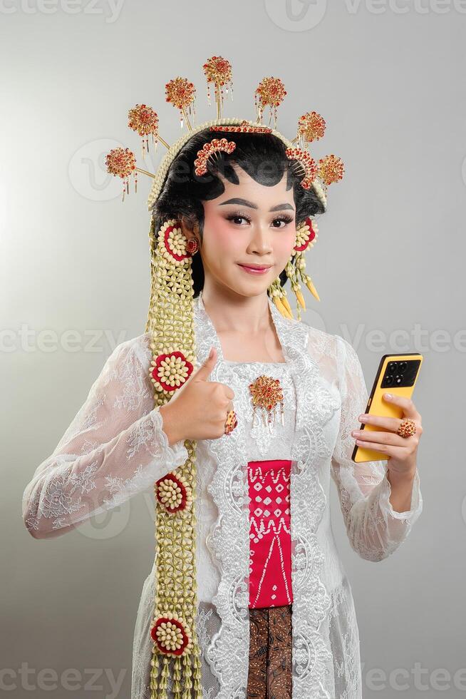 Indonesian bride in Solo Putri attire, wearing a golden headdress and jasmine garland. She smiles confidently, holding a smartphone and giving a thumbs-up, blending tradition and modernity. photo