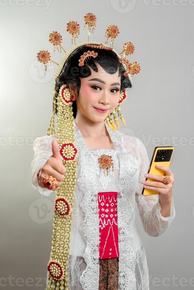 a Indonesian bride in Solo Putri attire, wearing a golden headdress and jasmine garland. She smiles confidently, holding a smartphone and giving a thumbs-up, blending tradition and modernity photo