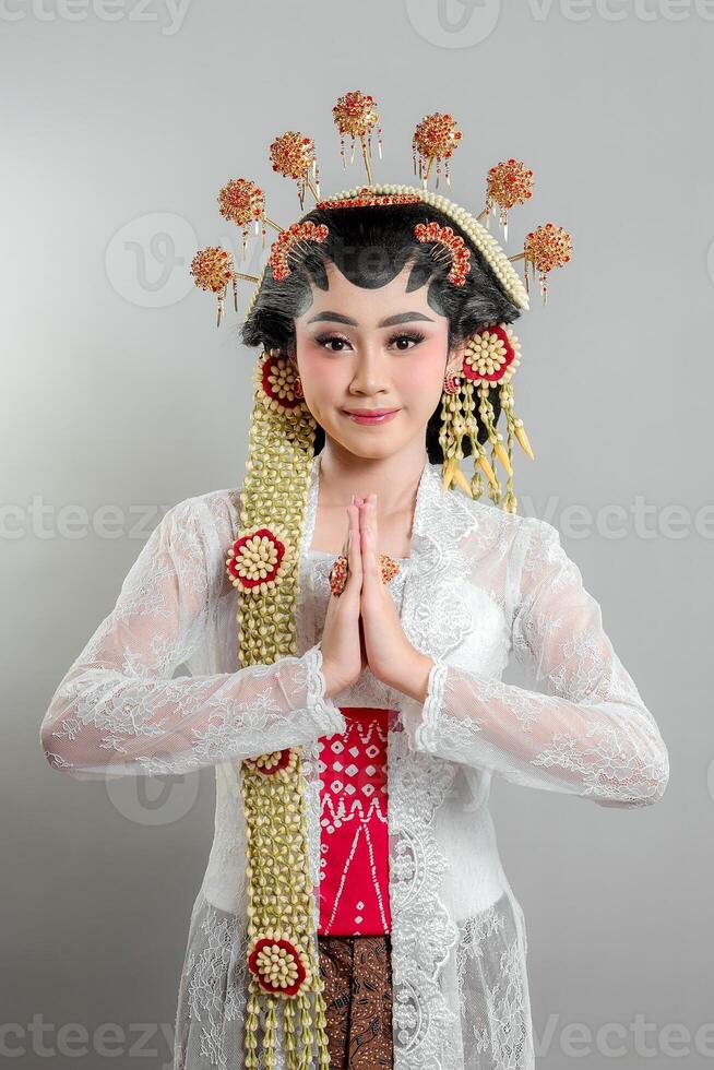 A Indonesian bride in Solo Putri attire stands gracefully with a soft smile hands in a respectful greeting. She wears a golden headdress, jasmine garland, and a white lace kebaya. photo