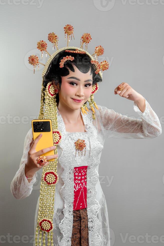 Indonesia bride in Solo Putri attire, wearing a golden headdress and jasmine garland. She confidently poses with a raised fist and holds a yellow smartphone, blending tradition with modernity. photo