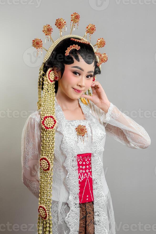 Indonesian bride in Solo Putri attire, wearing an intricate golden headdress and lace kebaya. She gently touches her ear while smiling softly, radiating elegance and grace. photo