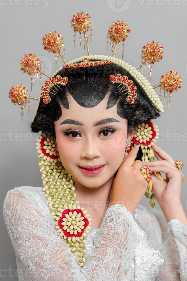 Indonesian bride in Solo Putri traditional attire, adjusting her golden earrings. She wears an ornate headdress, jasmine accessories, and a lace kebaya, exuding elegance and charm. photo
