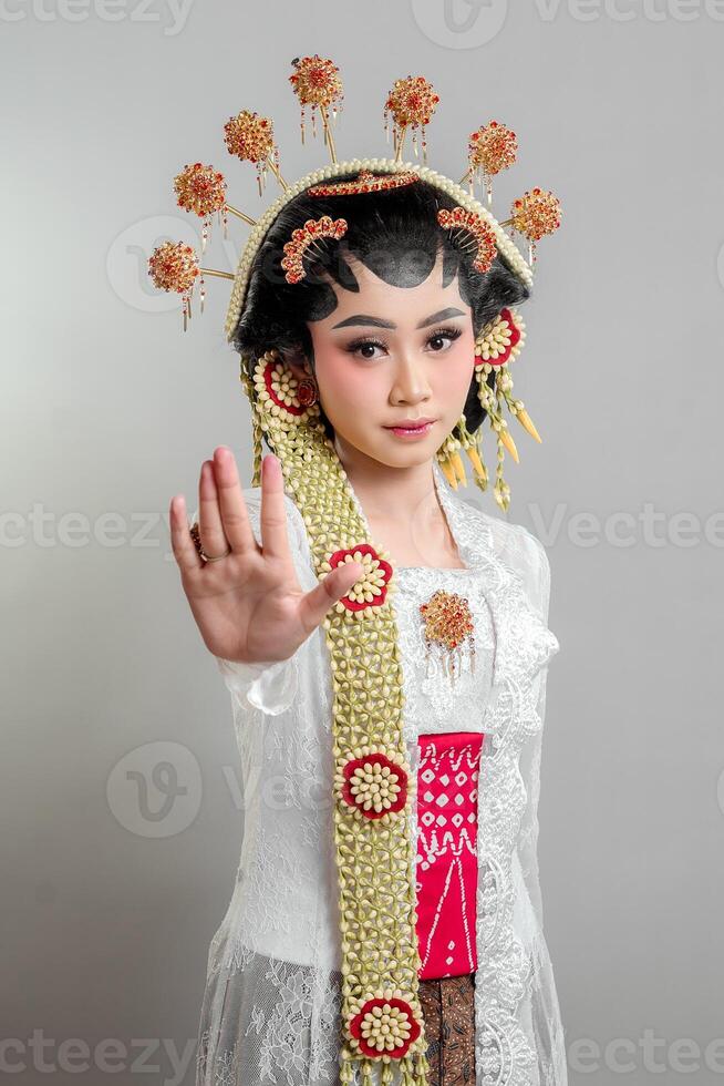 a Indonesian bride in Solo Putri attire, wearing a lace kebaya and ornate headdress. She gazes confidently, extending her hand in a stop gesture, exuding elegance and grace. photo