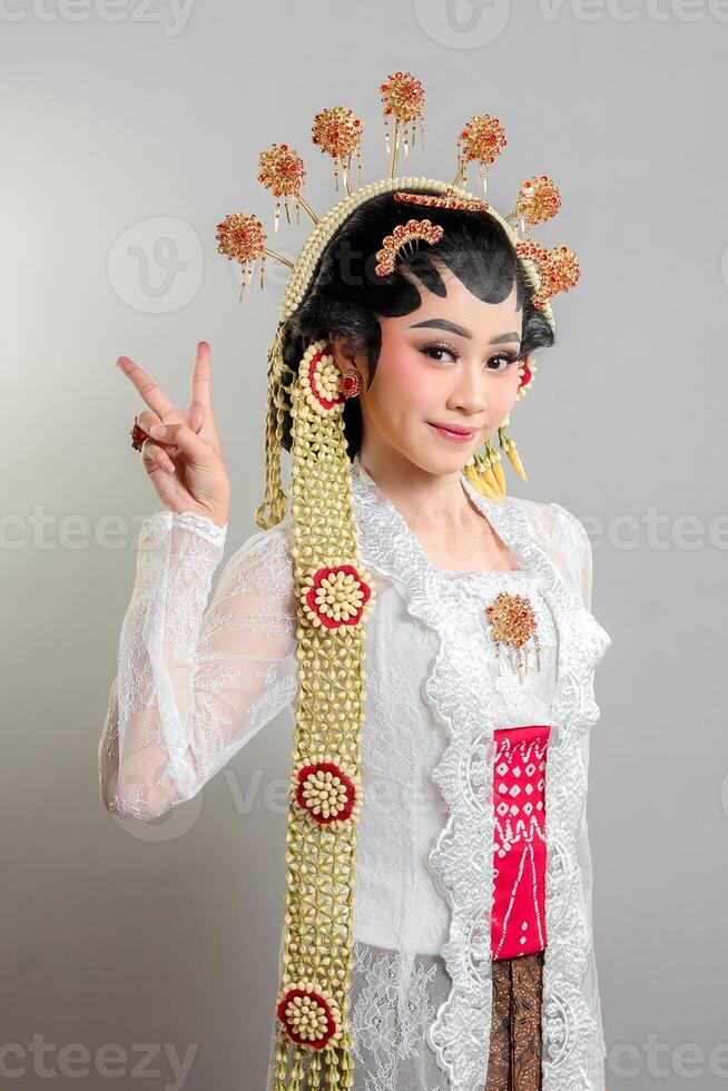 a Indonesian bride in Solo Putri attire, wearing a golden headdress and lace kebaya. She smiles gracefully while making a peace sign, exuding charm, confidence, and cultural elegance. photo