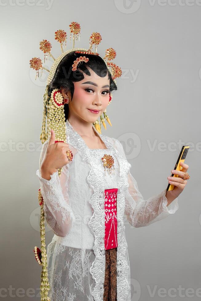 An Indonesian bride in Solo Putri attire smiles confidently while holding a smartphone and giving a thumbs-up. She wears an ornate golden headdress, elegant lace kebaya, and intricate jewelry. photo