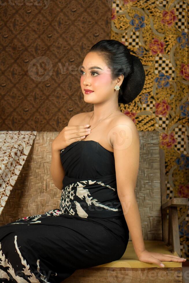 Indonesian woman wearing traditional batik fabric, sitting on a wooden bench. Vintage ethnic backdrop with rich patterns, cultural heritage, and graceful beauty in classic style. photo