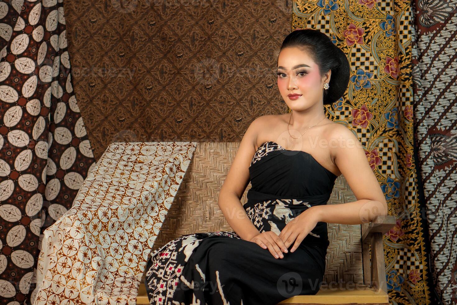 Indonesian woman in a black batik dress, sitting elegantly on a wooden bench. She exudes grace with a traditional hairstyle, vintage makeup, and cultural charm in a classic setting. photo