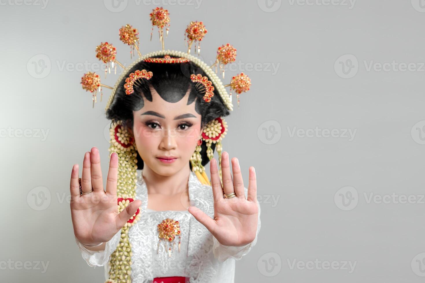 An Indonesian bride in Solo Putri attire, wearing an ornate headdress and lace kebaya. She raises both hands in a stop gesture with a serious expression, exuding elegance and grace. photo
