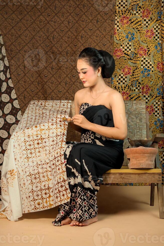 An Indonesian young woman in traditional attire making batik using a canting. She sits gracefully on a wooden bench, surrounded by handcrafted batik fabrics, showcasing cultural heritage photo