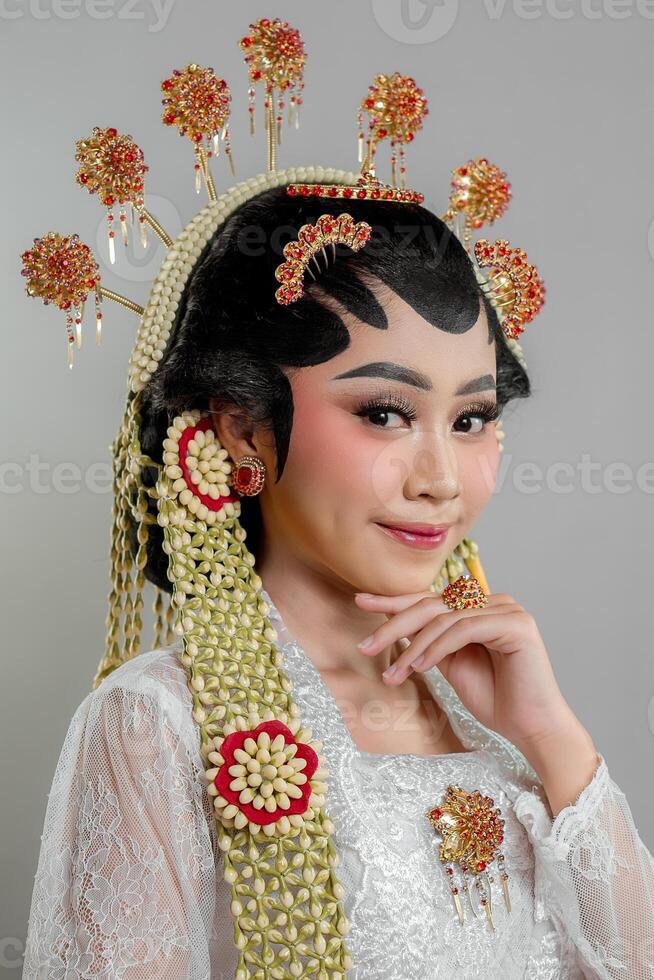 Indonesian bride in Solo Putri attire, wearing an intricate golden and jasmine headdress, white lace kebaya, and red jewelry, smiling gracefully with a confident expression. photo