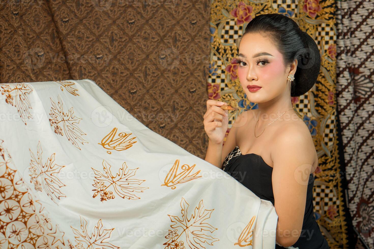 An elegant Asian woman in traditional attire holds a canting while making batik. She looks confident and graceful, surrounded by intricate batik patterns in a cultural setting. photo