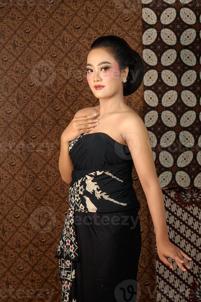 Elegant Indonesian woman in a black batik dress, posing gracefully with traditional Javanese style, against a patterned batik background. Cultural fashion portrait. photo