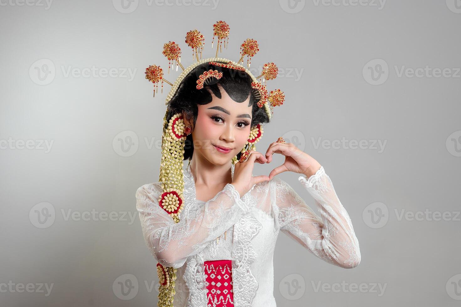An Indonesian bride in Solo Putri attire, wearing a lace kebaya, intricate headdress, and jasmine garlands. She smiles sweetly while forming a heart shape with her hands. photo