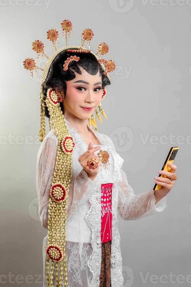 Indonesian bride in Solo Putri attire smiles while holding a smartphone. She wears an elegant golden headdress, intricate jewelry, a white lace kebaya, and traditional Javanese accessories photo