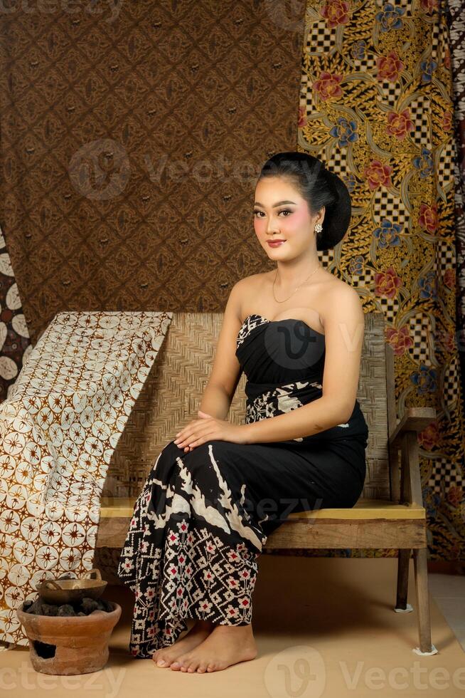Beautiful Indonesian woman in traditional batik dress, sitting gracefully on a wooden bench. Classic cultural setting with ethnic fabric, elegant makeup, and warm expression. photo