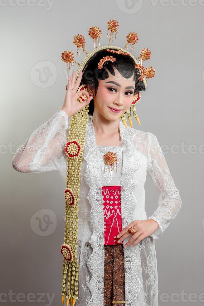 A Indonesian bride in Solo Putri attire wearing a lace kebaya, intricate headdress, and jasmine garlands. She smiles gracefully while making an OK hand gesture. photo