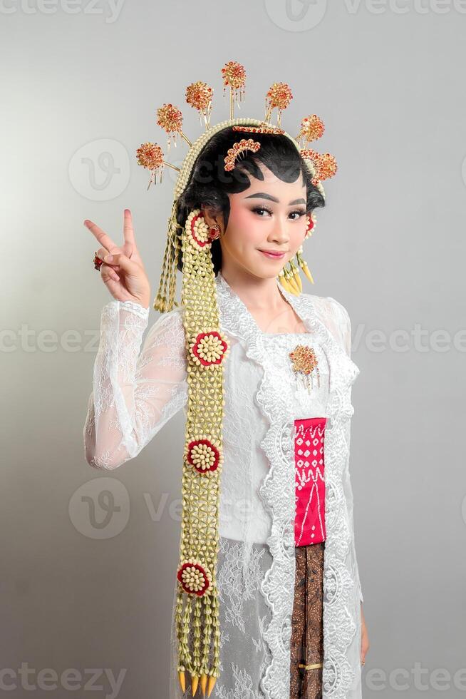 An Indonesian bride in Solo Putri attire, wearing a golden headdress and lace kebaya. She smiles gracefully while making a peace sign, exuding charm, confidence, and cultural elegance. photo