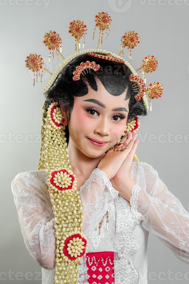 A Indonesian bride in Solo Putri traditional attire poses gracefully. She wears an intricate golden headdress, jasmine accessories, and a white lace kebaya, showcasing elegance with a soft smile. photo