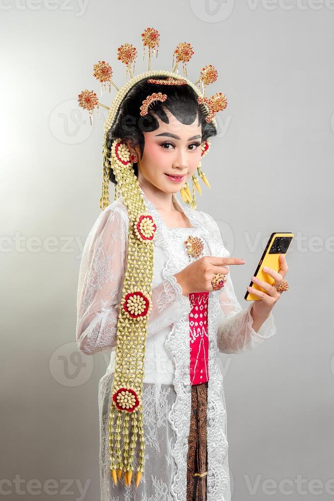 An Indonesian bride in Solo Putri attire smiles while using a smartphone. She wears a golden headdress, intricate jewelry, a white lace kebaya, and traditional Javanese accessories. photo