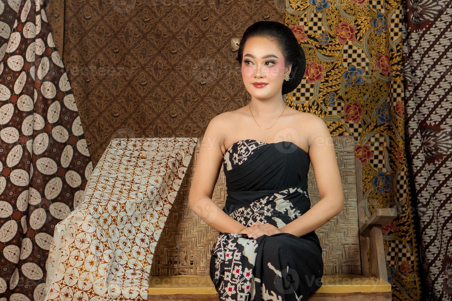 an Elegant Indonesian woman wearing a traditional black batik dress, sitting gracefully on a wooden bench. She poses with confidence, classic makeup, and an authentic Javanese hairstyle. photo