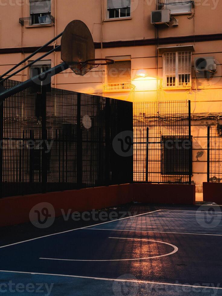 Basketball hoop and court glowing under street light at sunset, casting dramatic shadows in urban setting. Street light illuminating empty basketball court. highlighting court lines, hoop silhouette photo