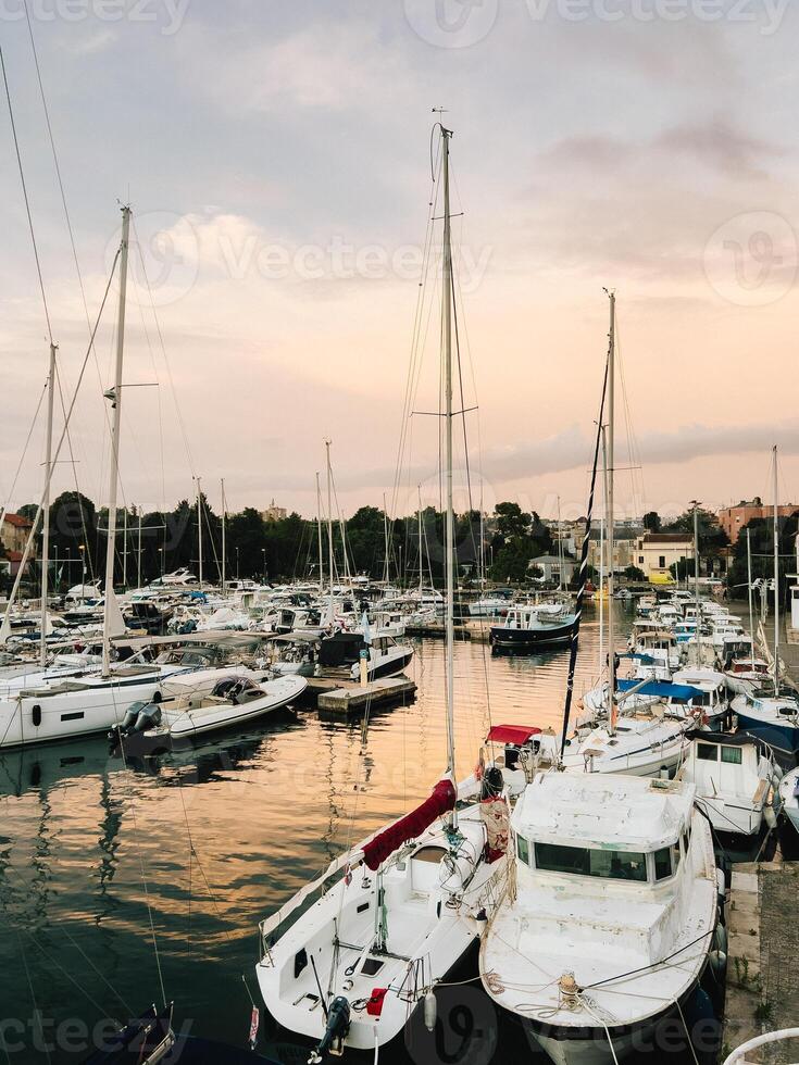 Sailboats and yachts are moored in tranquil marina as sun sets, casting warm golden glow on water and creating picturesque and serene scene of nautical beauty. Sailboats rest in marina during sunset photo