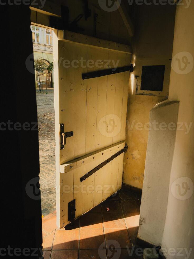 Sunlight streams into dark interior through an open, weathered wooden door, revealing glimpse of cobblestone street and building beyond, creating contrast between light and shadow. Wooden doorway photo