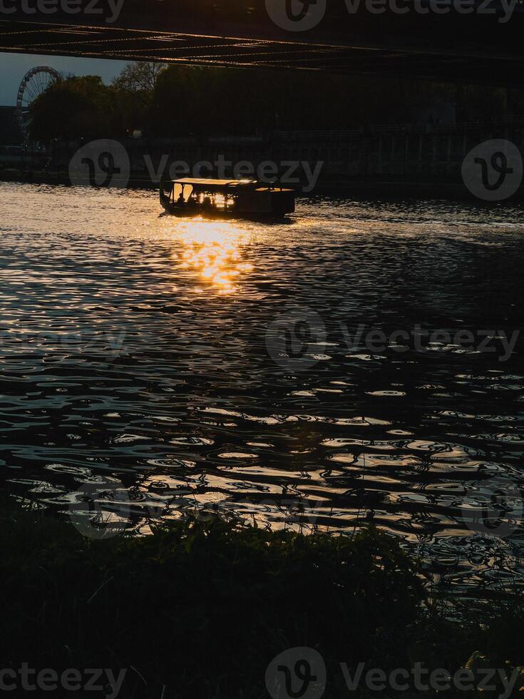Sightseeing boat transporting tourists on river during golden sunset, with Ferris wheel and bridge in background, creating romantic atmosphere. Golden sunset, Ferris wheel, historic bridge, cityscape photo