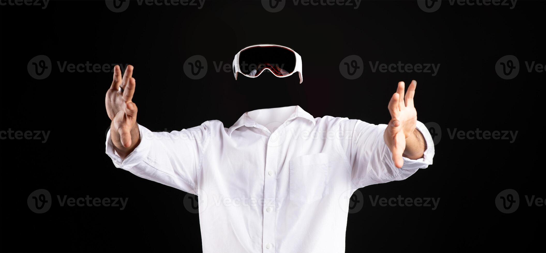 A user in a white shirt is immersed in virtual reality, wearing goggles while gesturing with hands. The background is completely dark, emphasizing the technology and user involvement. photo