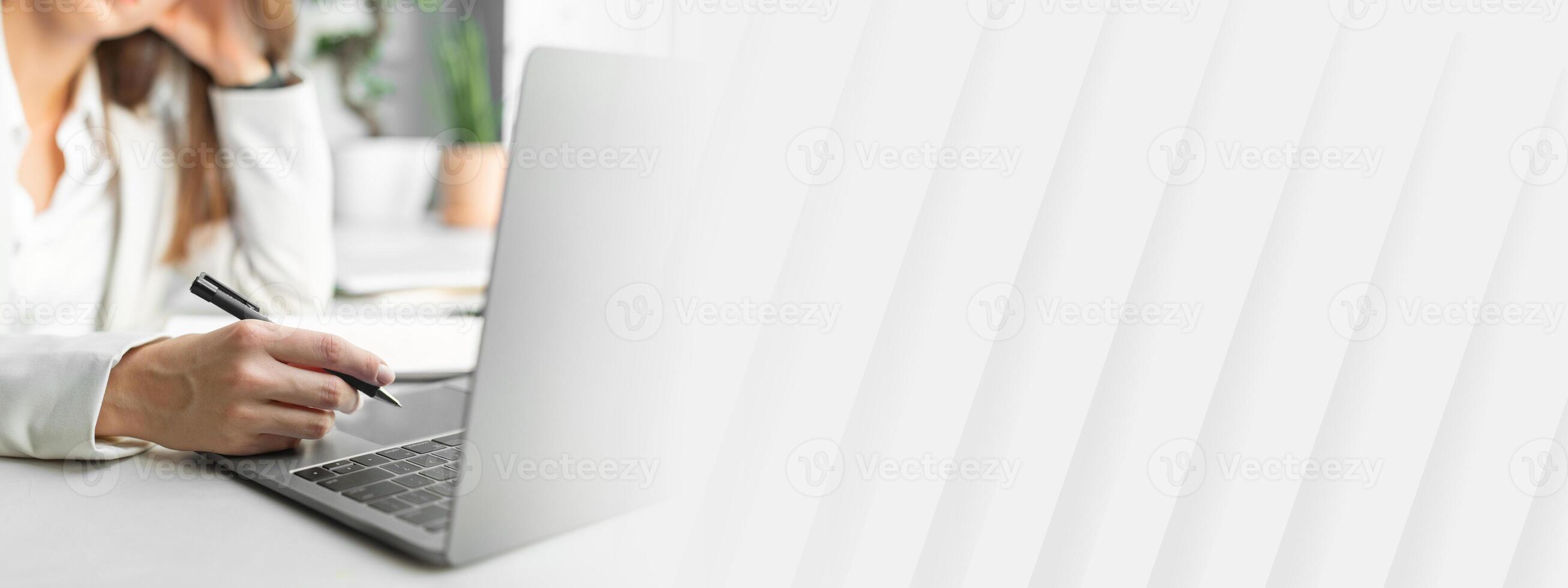 Pensive european businesswoman in formal wear holding pen, sitting in front of laptop in office interior, selective focus, closeup. Meeting remotely, work, call, business on gadget photo