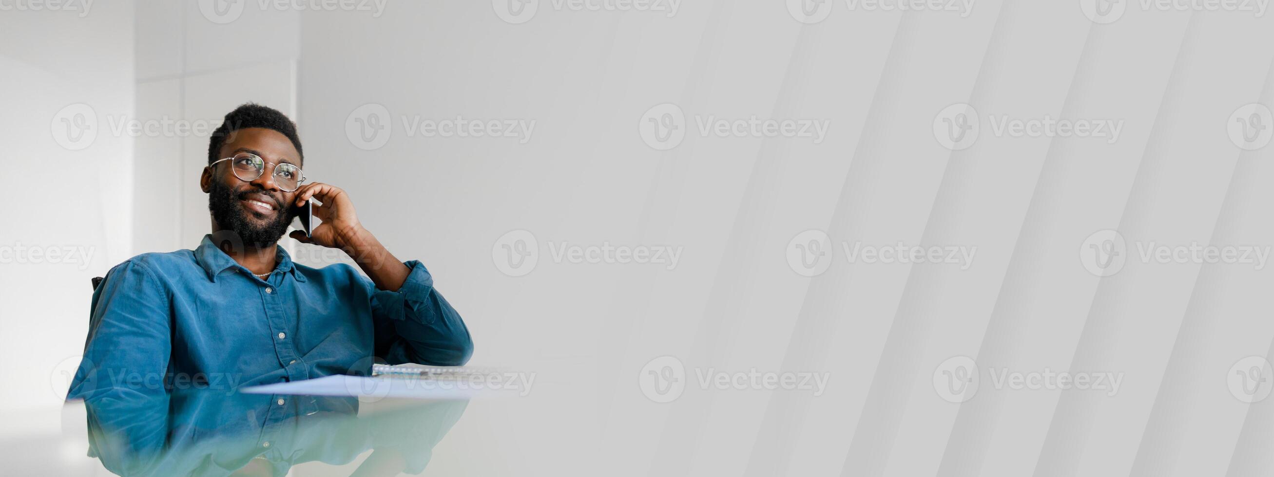 A young African American man sits in an office, smiling while talking on his phone. He appears confident and focused on his work, embodying a successful entrepreneurial lifestyle, copy space photo