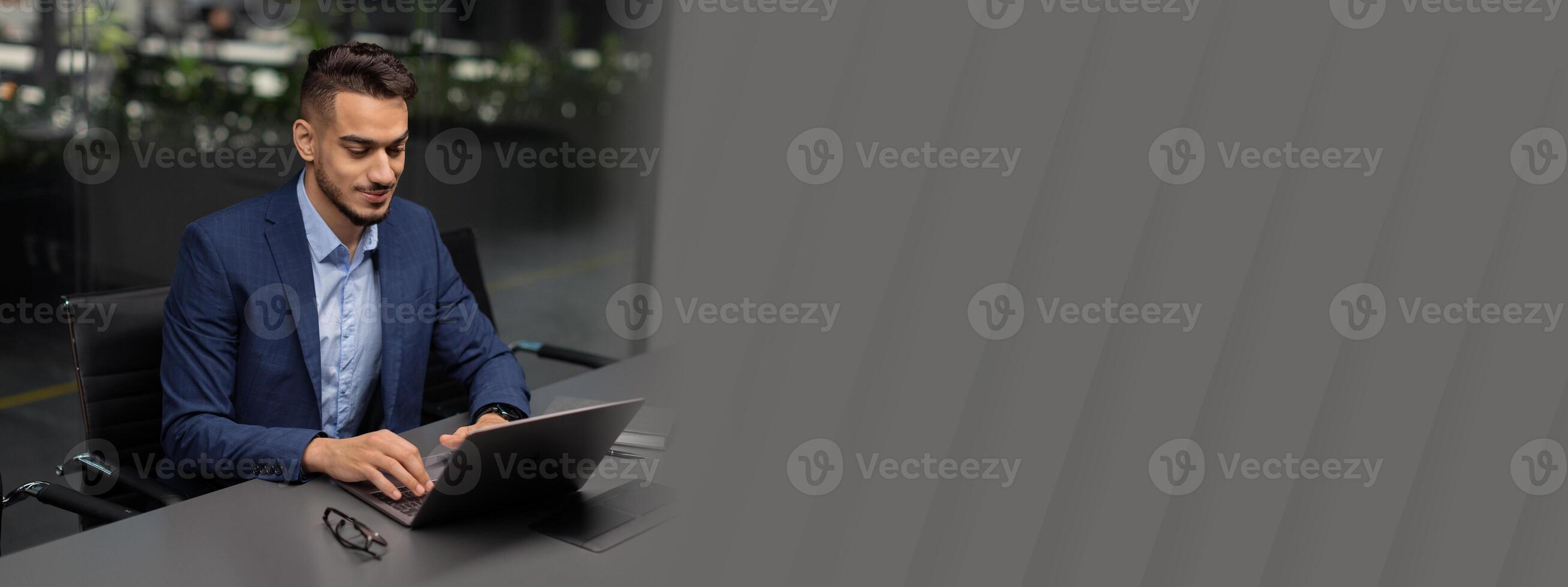 Handsome middle eastern entrepreneur working at modern office, arabic businessman sitting at workdesk, typing on laptop keyboard, drinking coffee, sending emails, copy space, high angle view photo