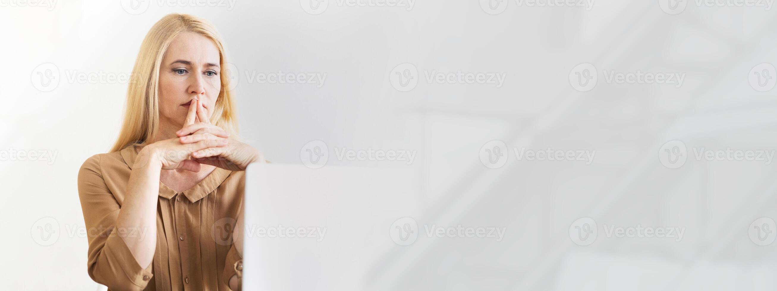 A woman sits at a desk in a modern workspace, deeply focused on her laptop. Sunlight filters through a window, illuminating her thoughtful expression as she contemplates her work, copy space photo