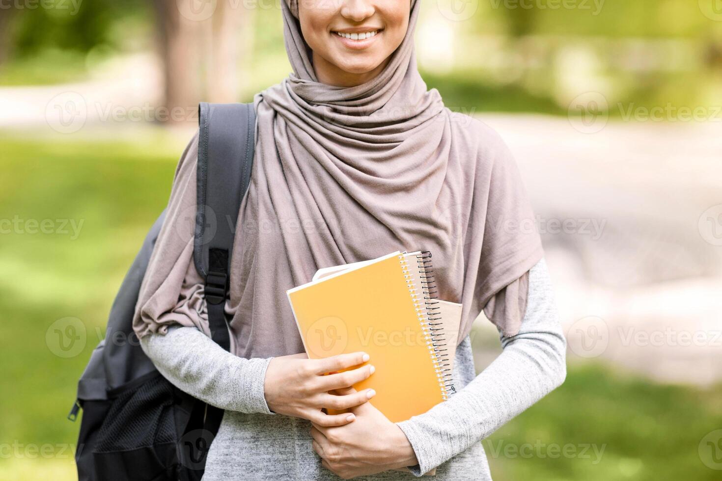 Pretty arab girl student walking by park after university, holding books, smiling at camera photo
