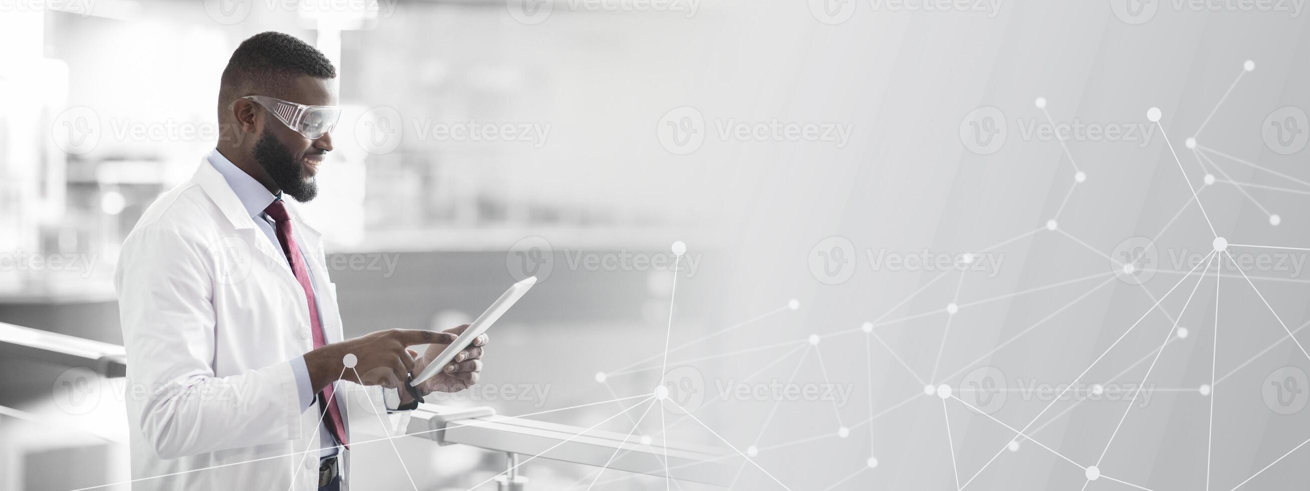 Cheerful african american guy in workwear and protective glasses medical scientist using digital tablet, analyzing data, posing at workplace, panorama with copy space. Jobs and occupations concept photo