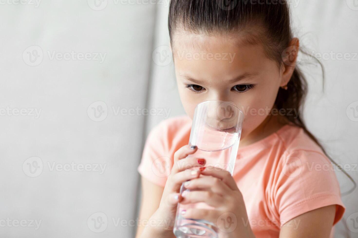 Japanese Kid Girl Drinking Water From Glass Sitting On Sofa At Home. Stay Hydrated Concept. Copy Space photo