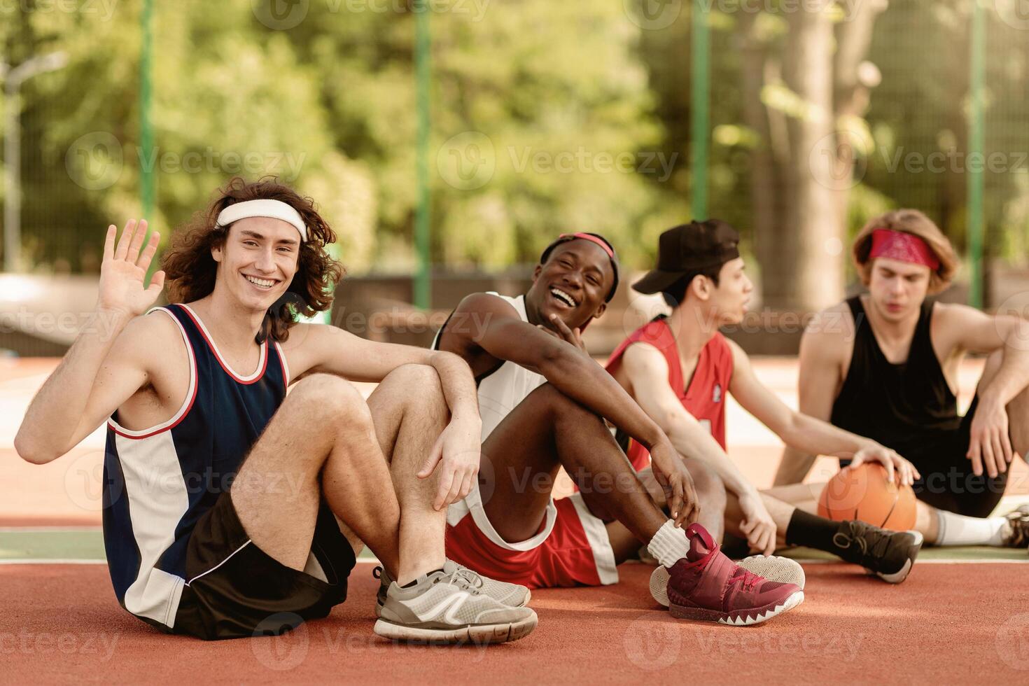 Happy Caucasian basketballer waving at camera and smiling, resting with his multiracial team after match at outdoor stadium, copy space. Portrait of professional sportsmen on break photo