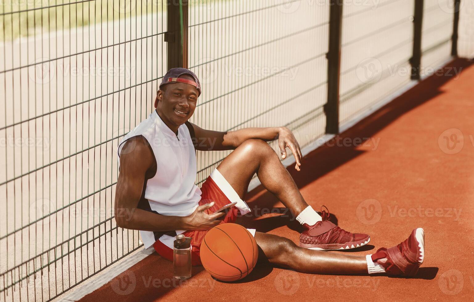 Handsome black sportsman with ball and mobile phone having rest at outdoor basketball court, panorama photo