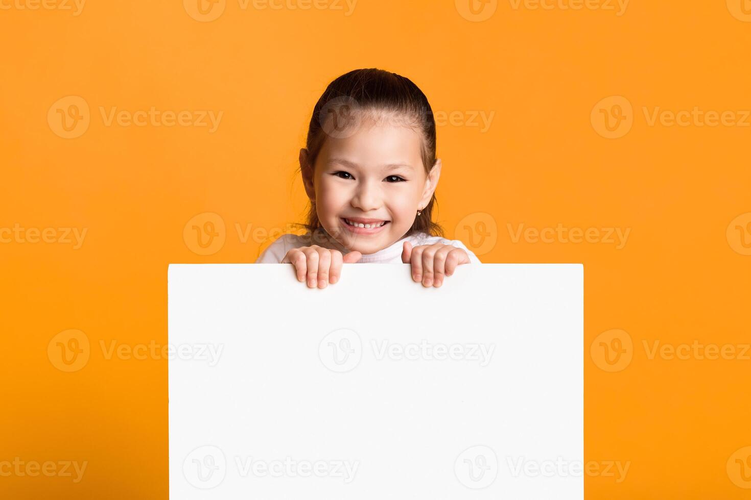 Advertisement Concept. Smiling little girl holding and looking out the square sign board with blank space, yellow wall photo